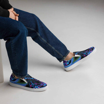 Men wearing slip on psychedelic canvas shoes with vibrant multicolor spiral design and white rubber sole sitting with blue jeans